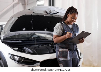 Black Woman Mechanic Engineer Holding Checklist Paper And Taking Notes On Clipboard, African Woman Is Standing Next To Car Engine Hood, Wearing Uniform Overalls. Side View Portrait
