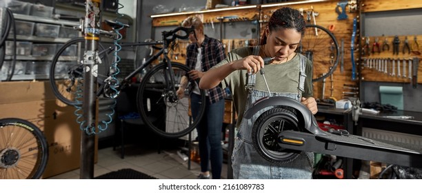 Black Woman Mechanic Fixing Electric Scooter With Screwdriver While Her Female Caucasian Colleague Installing Wheel Of Bicycle On Background In Modern Workshop. Bike Service, Repair And Upgrade