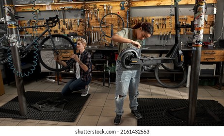 Black Woman Mechanic Fixing Electric Scooter With Screwdriver Near Female Caucasian Colleague Installing Wheel Of Bicycle With Wrench In Modern Workshop. Bike Service, Repair And Upgrade
