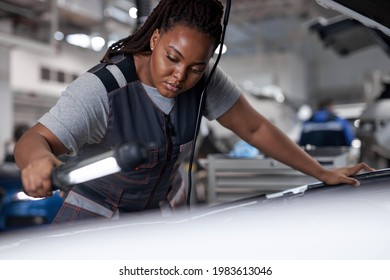 Black Woman Mechanic Looking To Car Engine And Holding Lamp, Car Master In Service Shop, Female Mechanic Repairer Service Technician Checks And Repairs The Engine Condition Under Hood Of Vehicle