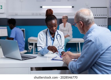 Black Woman With Medic Profession Looking At Documents With Elderly Patient Sitting At White Desk. African American Doctor And Old Man With Disease Writing On Paper Files For Checkup