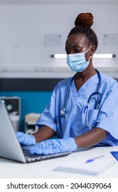 Black woman with nurse profession using modern laptop and technology while sitting at desk in medical cabinet. African american specialist with stethoscope wearing face mask for protection