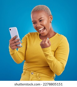 Black woman, phone and fist celebration in studio, blue background and winning online prize. Happy female model celebrate mobile promotion, bonus and excited for deal, success and competition winner