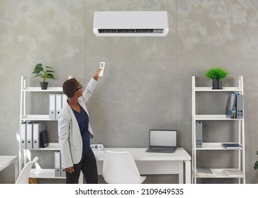 Black Woman Pointing A Remote Control At The AC On The Office Wall. Young African American Businesswoman Turns On The Air Conditioner To Enjoy Warm Air Or Cool Breeze In Her Workplace