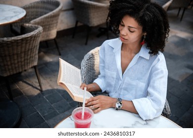 Black woman reading book and drinking juice on terrace table outdoors. View from above. Traditional no-technology hobbies.