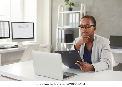 Black woman reading documents and thinking sitting at office desk with modern laptop. Business lady in glasses considering question, studying contract terms and conditions, making important decision