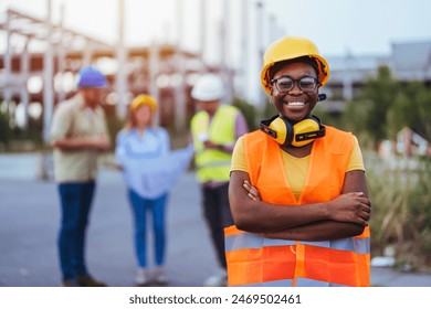Black woman in safety gear smiles at the camera with folded arms, her team engaged in discussion behind her at a busy construction site.