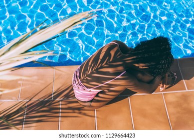 Black woman seating down relaxed in a swimming pool side with a palm tree shadow