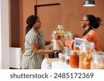 Black woman shops for fresh organic produce at local store. Customer purchasing nutritious bio food products from eco friendly shop, taking grocery bag from vendor at checkout counter.