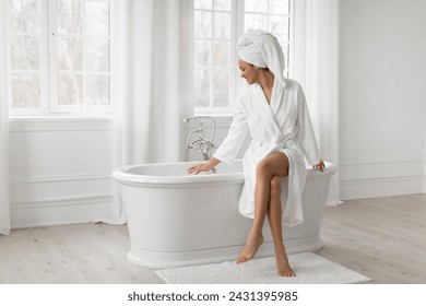 Black woman sitting on bathtub, wearing white bathrobe and towel on head, caring for body and skin in light bathroom at home, free space, full length - Powered by Shutterstock