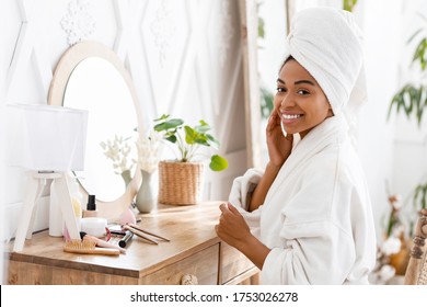 Black Woman Sitting At Toilet Table And Touching Her Perfect Skin, Wrapped In Bathrobe And Towel Pampering Herself After Bath, Looking At Camera - Powered by Shutterstock