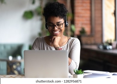 Black woman smart student girl sitting at table in university cafe alone wearing glasses looking at computer screen using headphones listening online lecture improve language skills having good mood