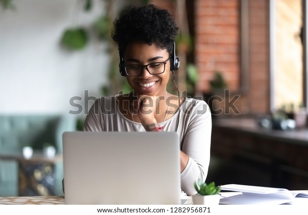 Black woman smart student girl sitting at table in\
university cafe alone wearing glasses looking at computer screen\
using headphones listening online lecture improve language skills\
having good mood