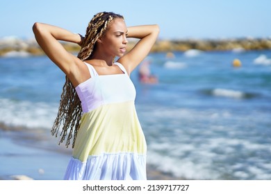 Black Woman In Summer Dress With Long Braids Holding Hands Behind Head And Looking Away While Relaxing On Beach Near Waving Sea On Sunny Day