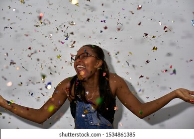 Black Woman Throwing Colored Glowing Confetti On A White Wall. 