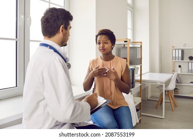 Black Woman Visiting Hospital. Female Patient Sitting On Couch In Exam Room And Talking To Professional Doctor About Chest Pain. General Practitioner Listening To Unhappy Young Lady Asking For Help