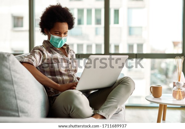 Black woman wearing face mask while\
working on a computer at home during virus epidemic.\
