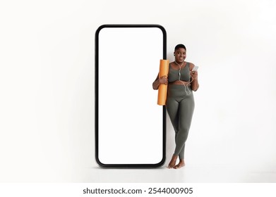 Black woman wearing green activewear holds a yoga mat and smartphone while leaning against a large blank phone silhouette. She radiates positivity, ready for fitness and wellness activities. - Powered by Shutterstock