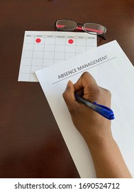 A Black Woman Writing On A Paper With The Words Absence Management On It. A Calendar Is In The Background. 