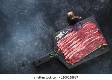 Black wooden chopping board with raw marbled beef bacon, horizontal shot with copyspace on a dark scratched metal background, top view