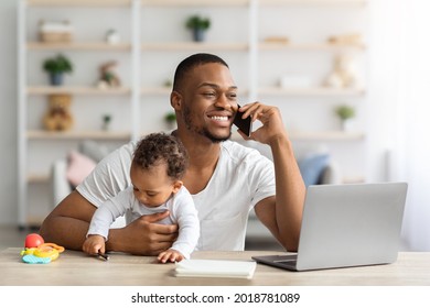 Black Working Father Talking On Cellphone While Babysitting His Infant Child At Home, Young Dad Sitting At Desk With Laptop Computer And Taking Care About Little Baby, Enjoying Paternity Leave