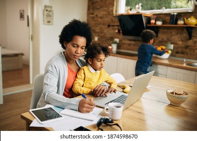 Black working mother taking notes while daughter is sitting on her lap and using laptop at home. Small boy is in the background. 