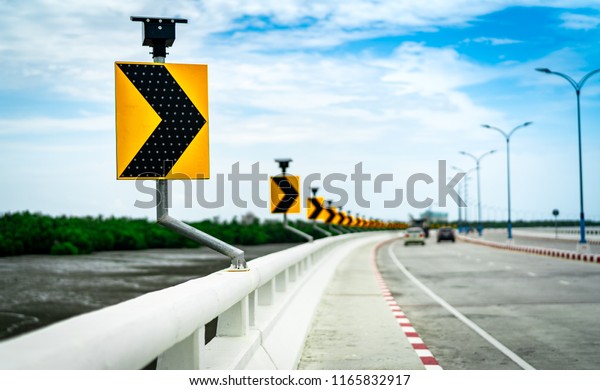 Black and yellow arrow on curve traffic sign on\
the bridge with solar cell panel ob blurred background of concrete\
road and car near mud flat and mangrove forest with beautiful blue\
sky and clouds.