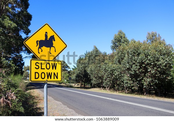 a black and yellow Slow Down, Horse\
Riders warning sign near a stretch of\
road