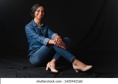 Black young beautiful happy smiling lady wearing jeans sitting on the floor with crossed legs and shoes against black background