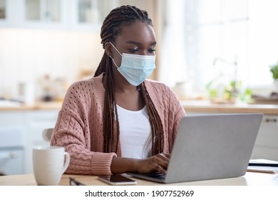 Black Young Business Woman Working From Home With Laptop, Wearing Protective Medical Face Mask During Coronavirus Quarantine Self-isolation, African Female Sitting At Table In Kitchen Using Computer