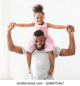 Black Young Happy Dad Carrying His Little Daughter On Shoulders, Posing At Home, White Background