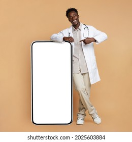 Black Young Male Doctor In Uniform Leaning And Pointing At Big Smartphone With Blank Screen, Showing Free Copy Space For Coronavirus Info Or Vaccination Campaign Design, Standing On Beige Background