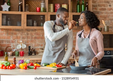 Black Young Man Feeding His Beautiful Woman While Cooking Together At Kitchen, Empty Space