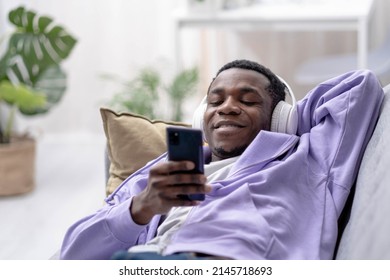 Black young man using smartphone at home lying on couch, pressing finger, reading social media internet, typing text or shopping online, Making video call