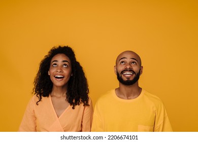 Black Young Man And Woman Looking Upward While Smiling Together Isolated Over Yellow Background