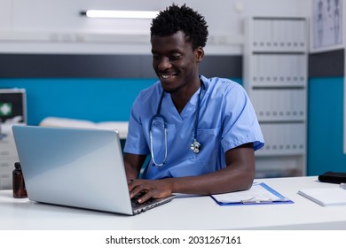 Black young man working as nurse at medical clinic wearing uniform and stethoscope. African american person sitting at desk while using modern laptop, typing on keyboard in cabinet