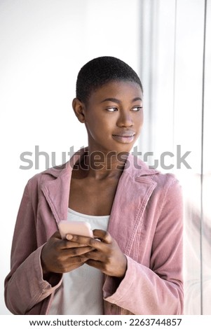 Black Young woman with buzz cut hairstyle and pink blazer texting on phone while leaning on glass window and looking away. vertical copy space