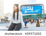 Black young woman portrait in Berlin. She is sitting on a concrete wall in Alexanderplatz with ubahn, the underground train, sign on background.