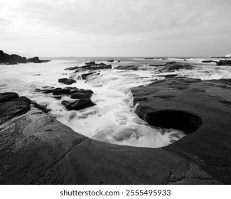 
A black-and-white coastal Papuma beach scene featuring smooth waves cascading over rocky formations, creating natural contours and textures under a cloudy sky. - Powered by Shutterstock