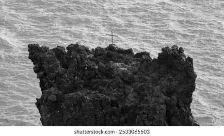 A black-and-white image of a rugged rocky cliff with a cross at the top, overlooking the ocean, creating a dramatic and isolated scene - Powered by Shutterstock