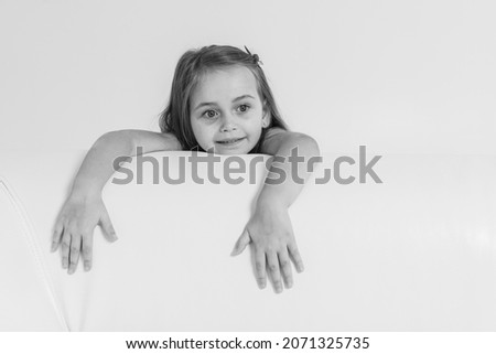 Black-and-white photo, a little blonde, cute emotional girl in a dress with blond hair leaned on the side of a white leather sofa on a white background, hung his hands, looks.
