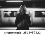 A black-and-white photo of a woman with a backpack standing on a subway platform, viewed from behind. A train blurs by, capturing a quiet moment of reflection in a fast-paced urban setting.