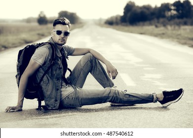 Black-and-white Portrait Of A Handsome Young Man Hitchhiker Sitting On A Highway. Adventure And Tourism Concept. Jeans Style.