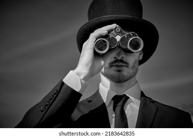 Black-and-white Portrait Of A Man In A Black Suit And Top Hat Looking Into The Distance Through Binoculars. Surrealist Style.