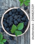 Blackberries with leaves in a ceramic bowl on a wooden background.
