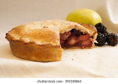 Blackberry Pie With Slice Missing On White Cloth And Blackberries And Apple In The Background.