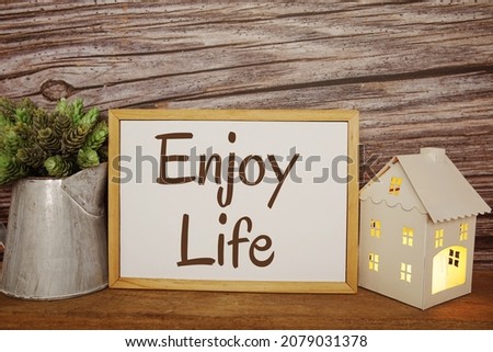 Blackboard with the text ENJOY LIFE decorate with artificial plant in watering plant and LED candle in house lantern on wooden background