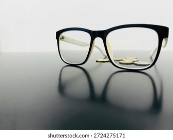 Black-framed eyeglasses and a small stack of silver and gold coins resting on a reflective dark surface against a plain white background.