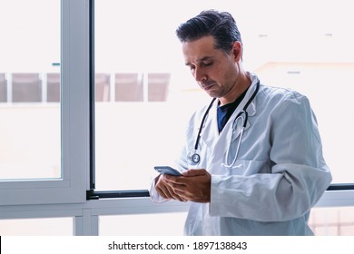 Black-haired Doctor In White Coat And Stethoscope Looking At His Mobile Phone, In A Window. Medicine Concept