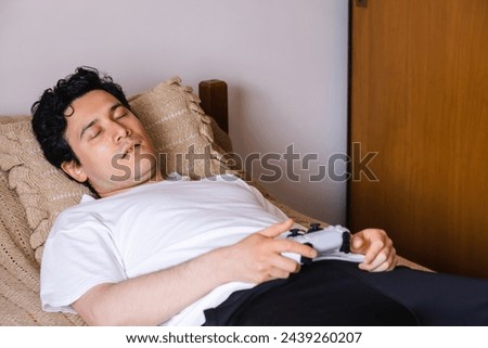 Black-haired Latin man falls asleep in his bed while he plays video games, horizontal photo